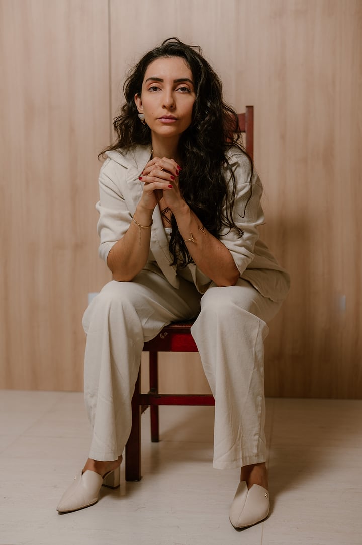 Ana Luiza sentada numa cadeira, de conjunto claro, com as mãos entrelaçadas